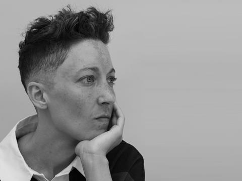 Black and White headshot of a white queer person with olive, freckled skin. Short dark fade, curly on top. Light eyes peer into the distance, as she rests her chin on her hand.